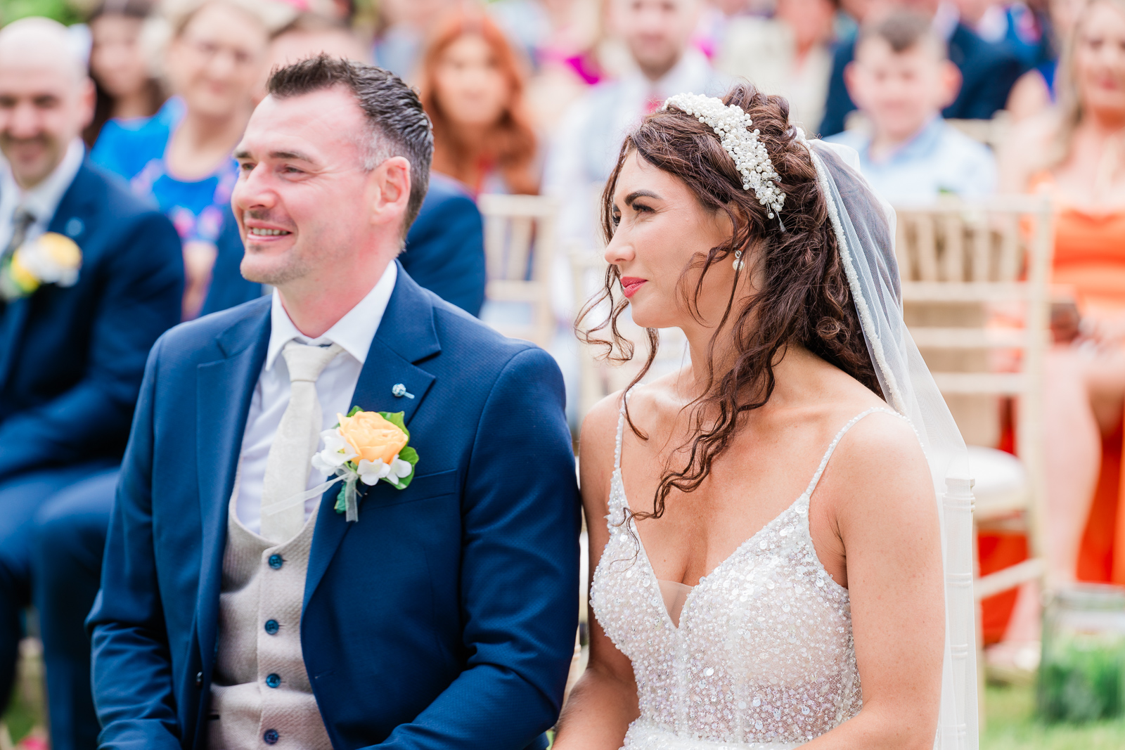 Claire and Anthony sitting and smiling during their wedding ceremony at Drumhalla House, Rathmullen. The bride is in a sparkly gown, and the groom is in a navy suit.